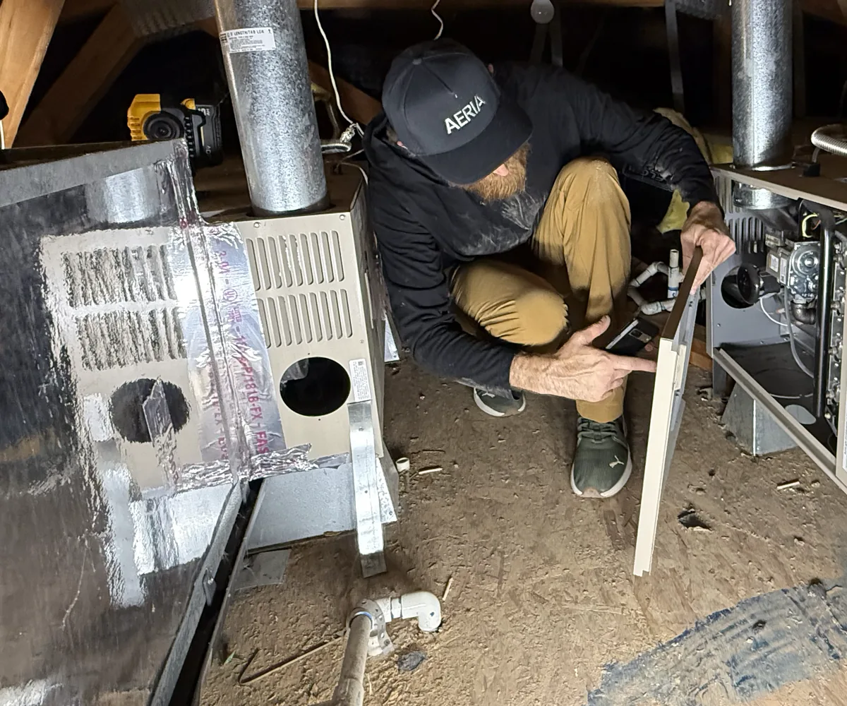 Technician in an AERIA cap crouched beside an attic furnace with its access panel removed